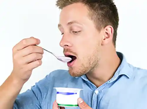 Young caucasian man wearing a blue button down shirt eating a spoonful of yogurt out of a small container.
