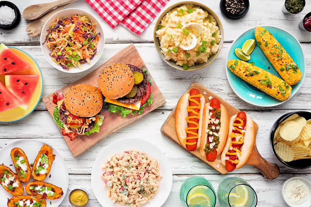 Assorted summer picnic foods including burgers, hot dogs, salads, watermelon, and chips arranged on a rustic white wooden table for outdoor dining.