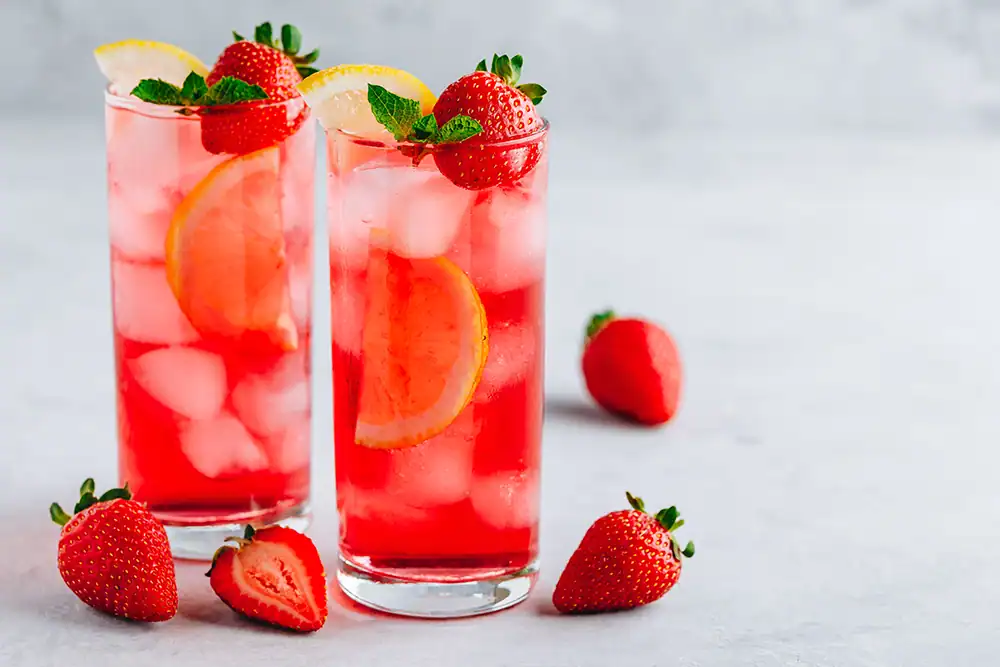 Refreshing Strawberry Lemonade Drinks Two tall glasses of strawberry lemonade with fresh strawberries, lemon slices, and mint leaves on a light background.